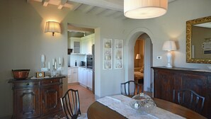 A dining area with a wooden table, chairs, and a chandelier. There is a kitchen in the background with white cabinets and a stove. - Holiday Home in Cortona With Pool & Valley Views (Cortona)