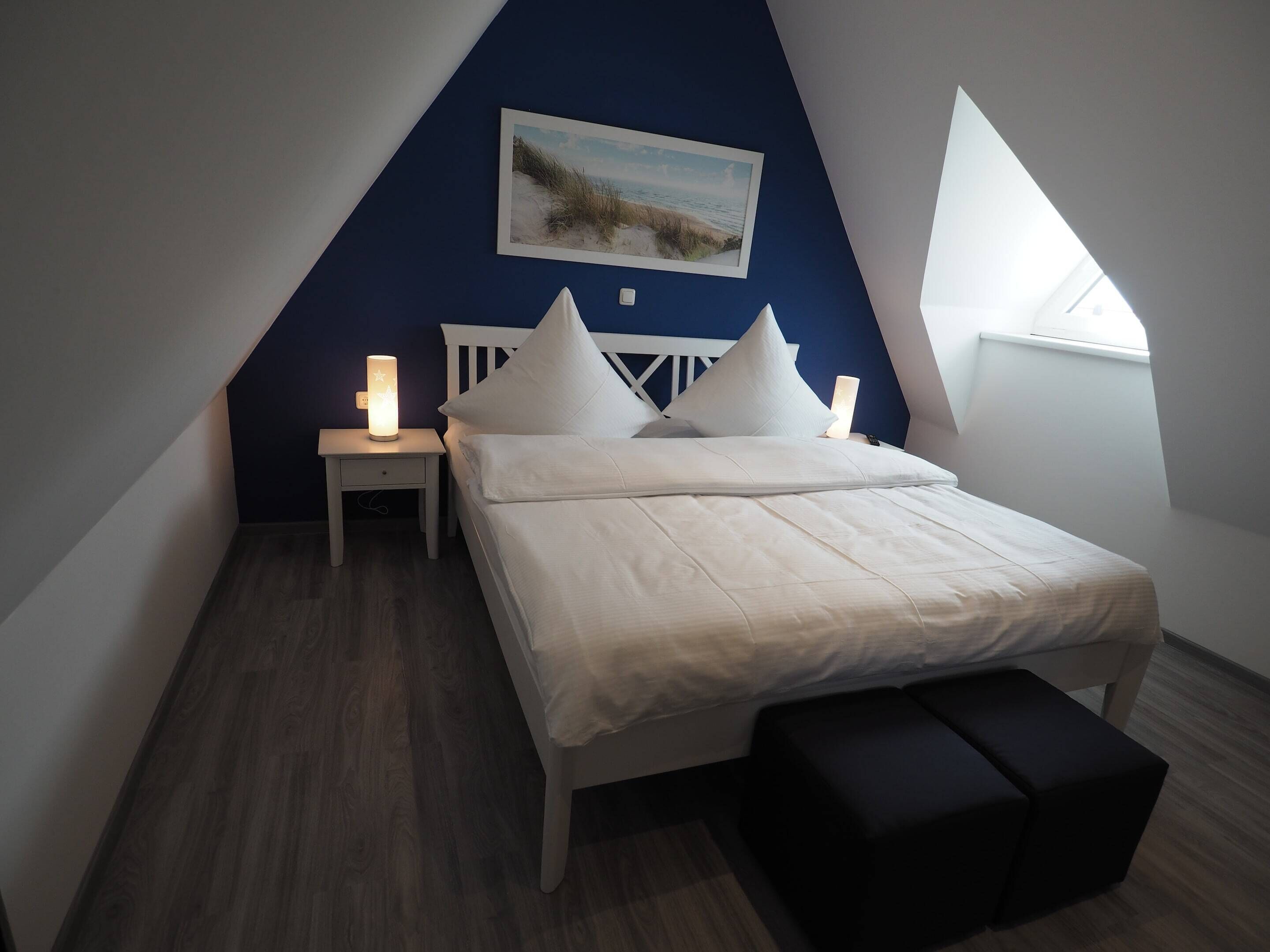 A bedroom with a sloped ceiling, a bed with white bedding, a nightstand with a lamp, a painting on the wall, and a window.