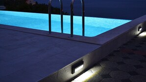 Une piscine dotée de deux rampes métalliques, d’une terrasse en pierre et offrant une vue sur la mer et les montagnes en arrière-plan.