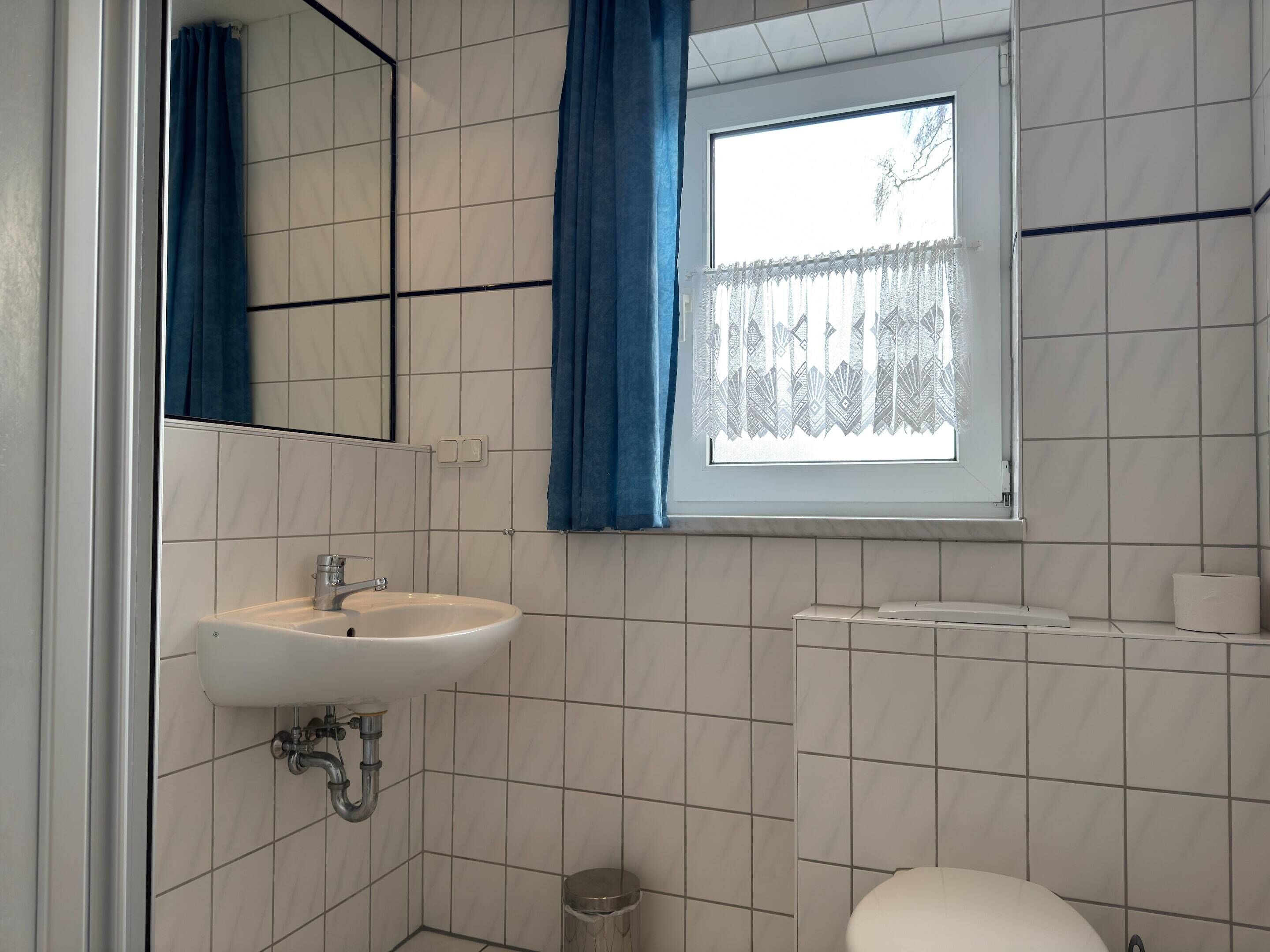 Ein Badezimmer mit einem weißen Waschbecken, einem Spiegel, einem Fenster mit blauen Vorhängen und einer Toilette.