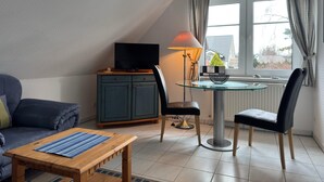 A cozy living room with a sofa, a coffee table, a TV on a cabinet, a lamp, and a window with curtains. - Ferienwohnung in Zingst (Zingst)