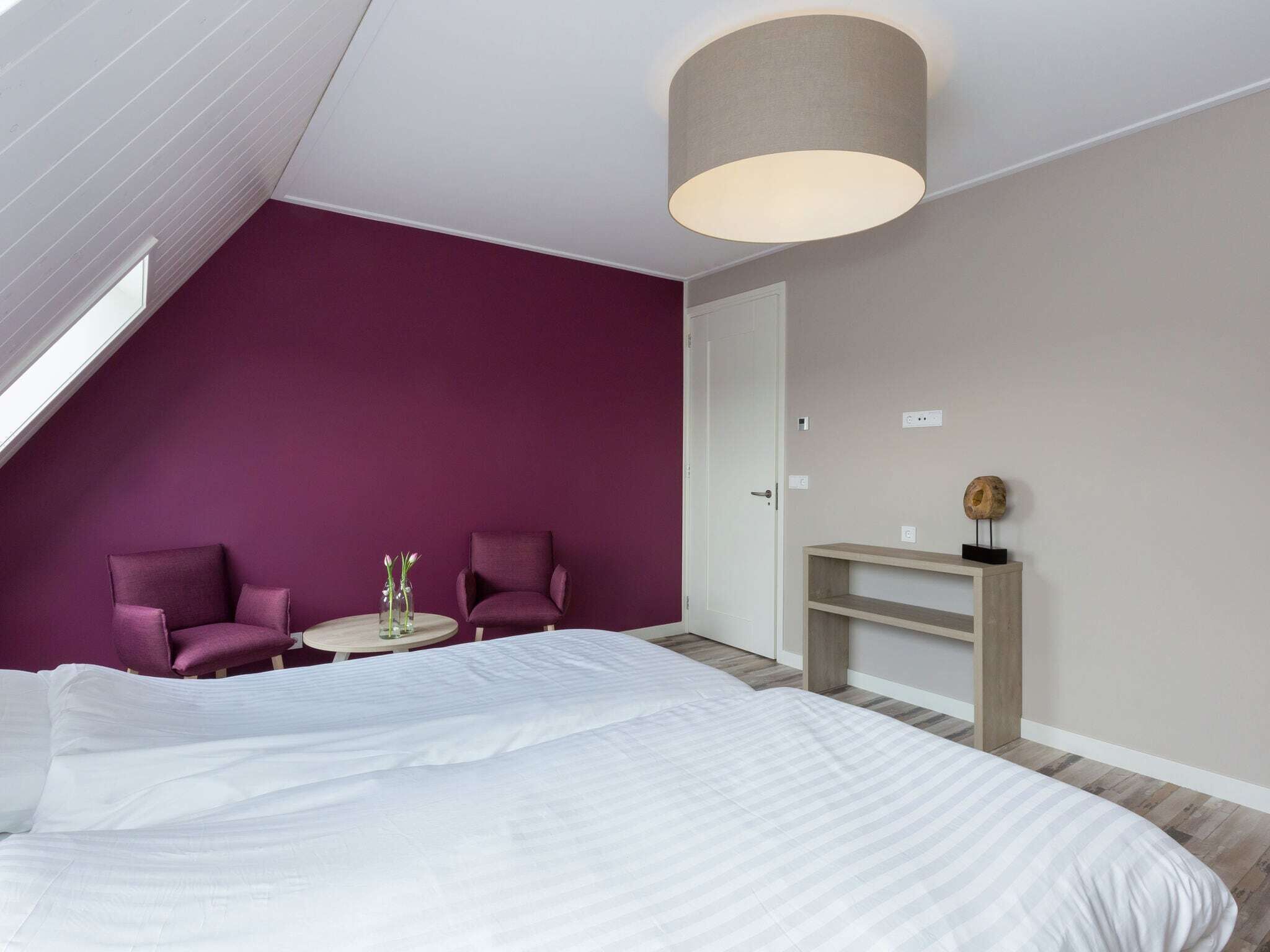 A bedroom with a large bed, two chairs, a small table with flowers, a wooden console, and a sloped ceiling with a light fixture.