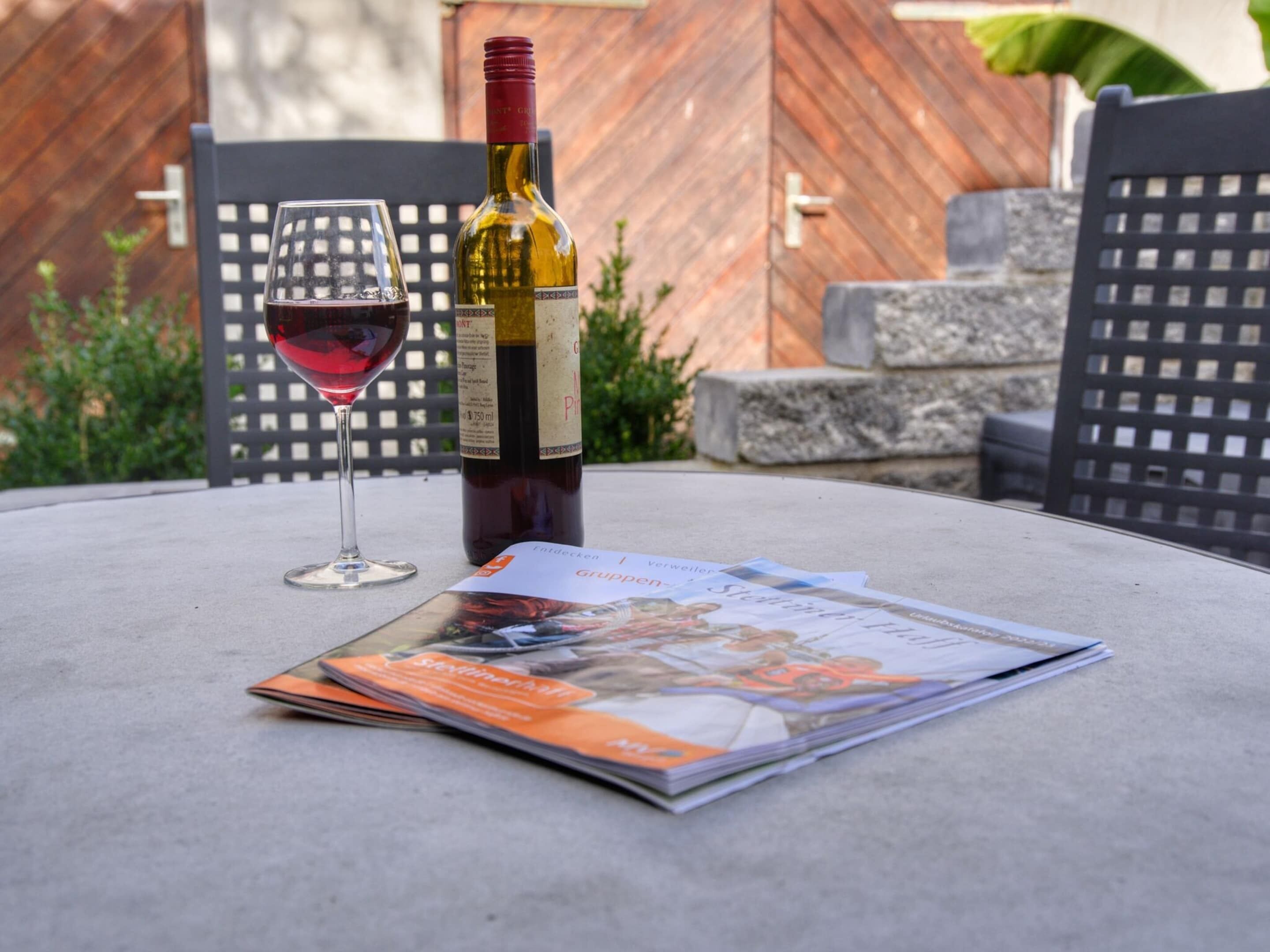 Una botella de vino, una copa de vino tinto y una revista sobre una mesa al aire libre.