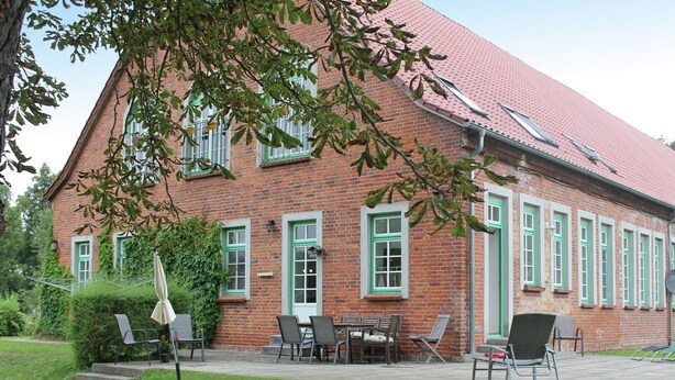 Ein Backsteinhaus mit grünen Fensterrahmen, einem roten Dach und einer Terrasse mit Gartenmöbeln.