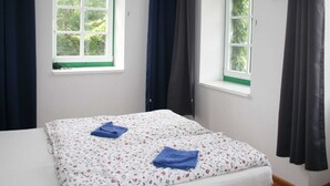Ein Zimmer mit zwei Betten, jeweils mit einem blumigen Bettüberwurf und einem blauen Handtuch. Zwei Fenster mit weißen Rahmen und grünen Fensterbänken sowie dunkle Vorhänge.