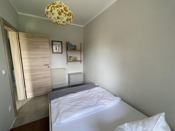 Un dormitorio con cama, armario, radiador, una mesita y estantería con libros y un juguete.
