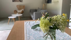 A living room with a wooden table, a glass vase with flowers, a patterned tablecloth, a grey sofa, a small round table, and a chair. - Apartment Near Saaler Bodden Lagoon (Saal)