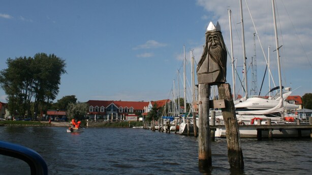 A marina with boats docked, a wooden post with a statue, and a building with a red roof in the background. - Bungalow in Tessmannsdorf Nahe Salzhaff (Am Salzhaff)