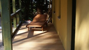 Una terraza de madera con un banco, rodeada por un muro amarillo y barandillas de madera, con árboles al fondo.
