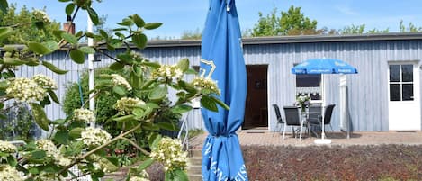 Ulkoterassi, jossa on pöytä ja tuoleja, sininen aurinkovarjo ja rakennus, jonka julkisivu on tehty puusta.