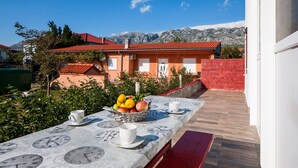 Une terrasse avec une table dressée pour deux personnes, agrémentée d’un panier de fruits et de tasses, d’où l’on peut admirer une montagne et un bâtiment au toit rouge.