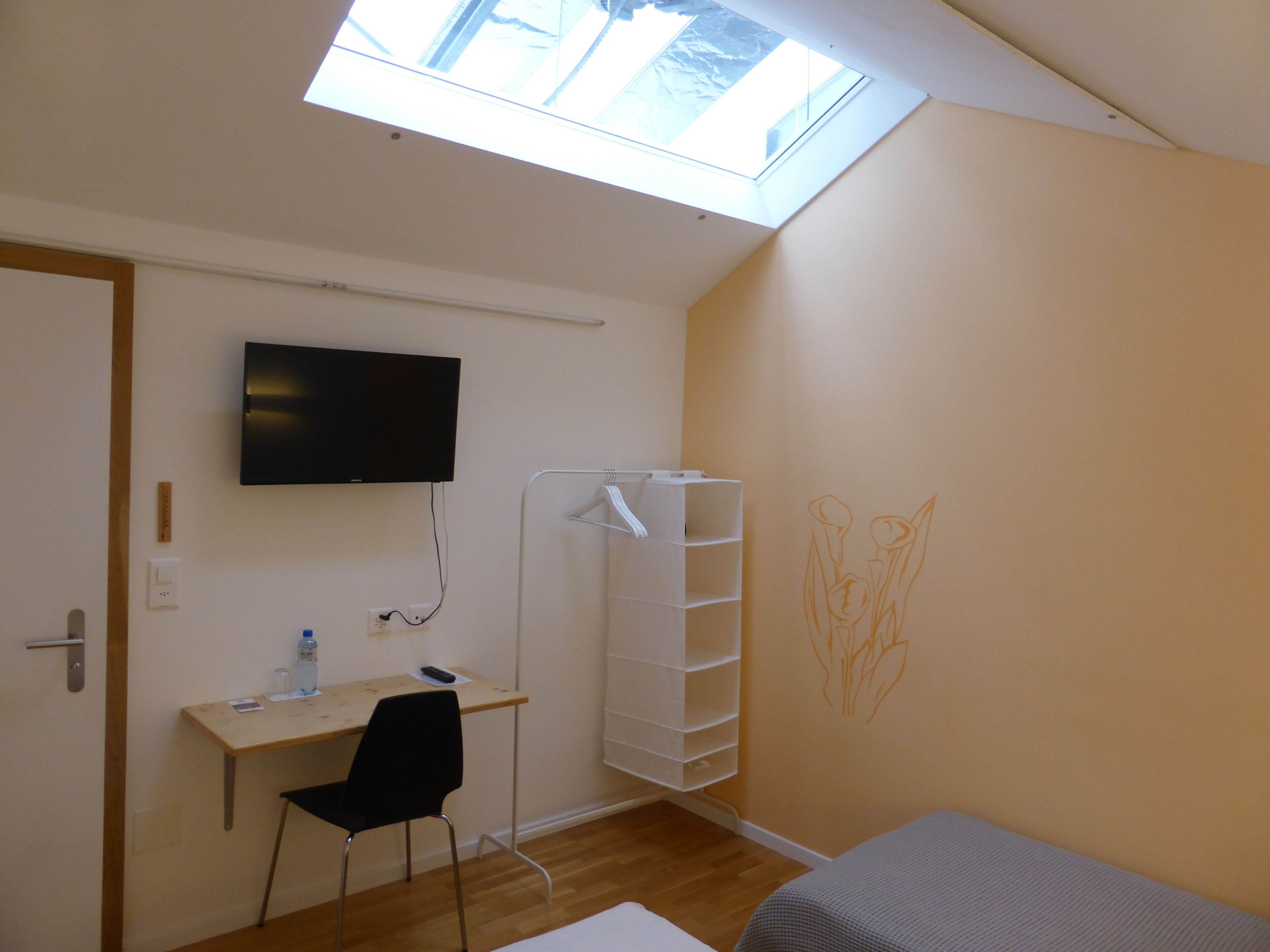 Ein Zimmer mit Dachfenster, Schreibtisch mit Stuhl, einem Bett und einem an der Wand befestigten Fernseher.