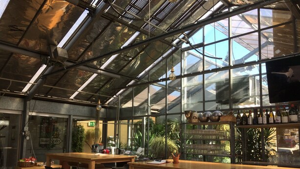 Un invernadero con mesas y sillas de madera, una zona de bar con botellas, y vistas a la vegetación exterior.