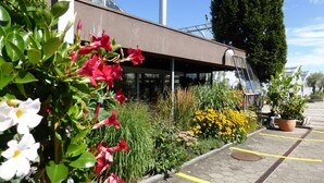 Un edificio con un área exterior cubierta, un estacionamiento con líneas amarillas y diversas plantas y flores.