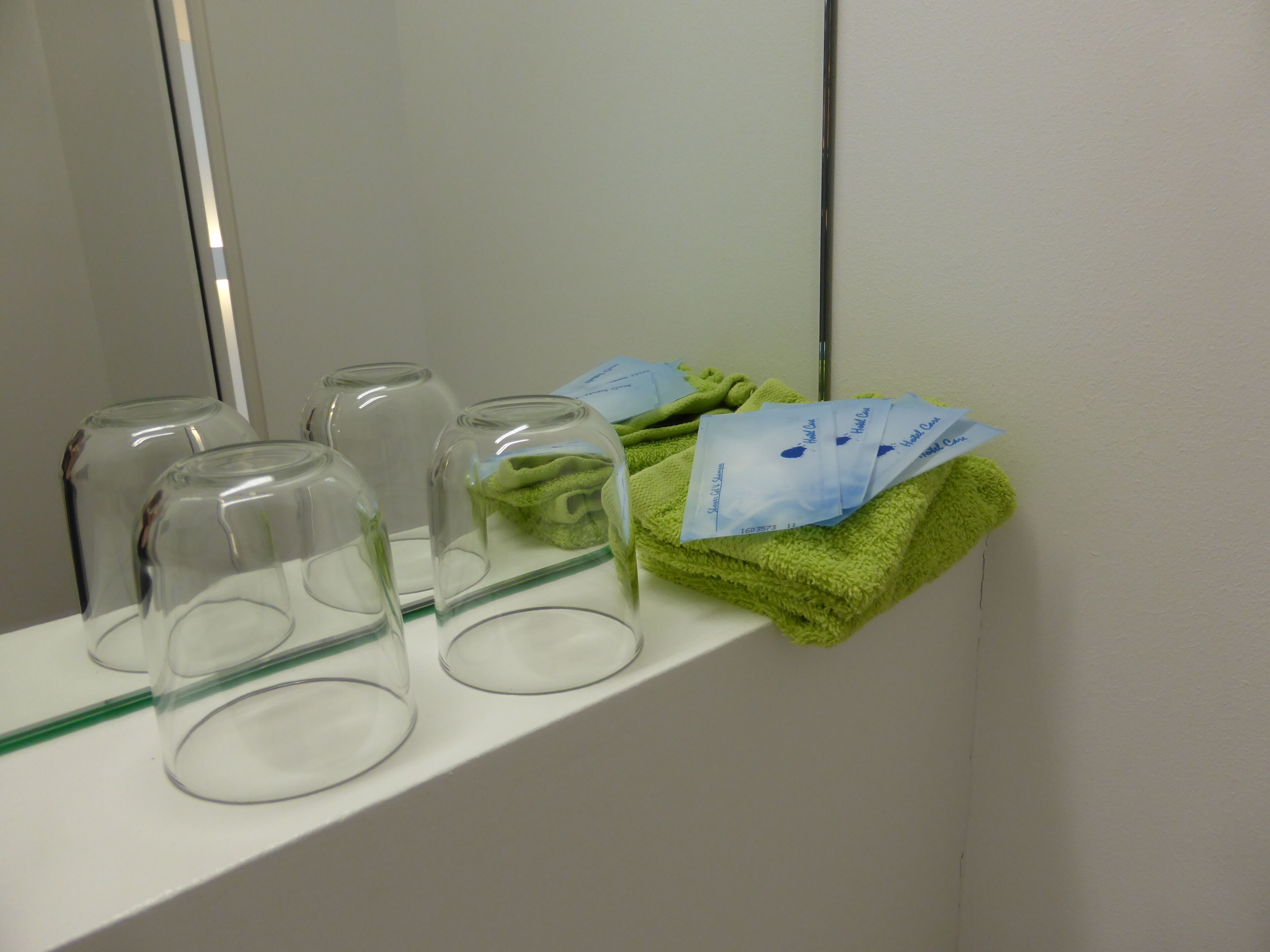 Un baño con espejo, repisa con vasos de vidrio y toalla verde.