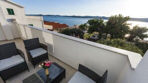 Un balcon doté de meubles en osier et d’une table en verre, offrant une vue sur une étendue d’eau et un paysage vallonné.