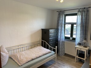 A bedroom with a bed, a dresser, a window with patterned curtains, a radiator, and a small table with a lamp.