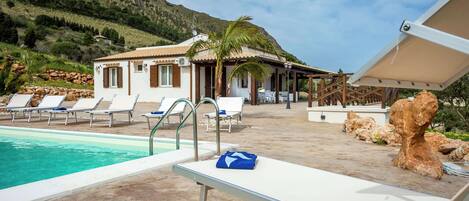 A pool area with lounge chairs, a small building, and a mountain backdrop.