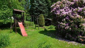 Un parque infantil con tobogán y columpios, rodeado de abundante vegetación y arbustos florecientes.