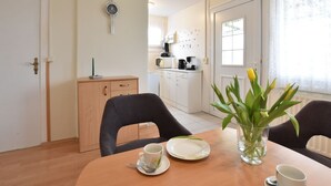 A dining area with a wooden table, chairs, a vase with flowers, and a kitchen in the background. - Strandbungalow, Boltenhagen (Boltenhagen)