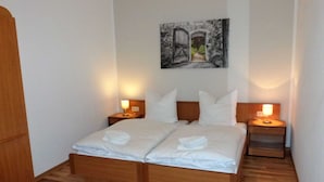 A neatly made double bed with white linens and two pillows, flanked by wooden nightstands with lamps, in a room with wooden flooring and a painting on the wall. - Geraeumige Ferienwohnung mit Terrasse in Kluetz (Klütz)