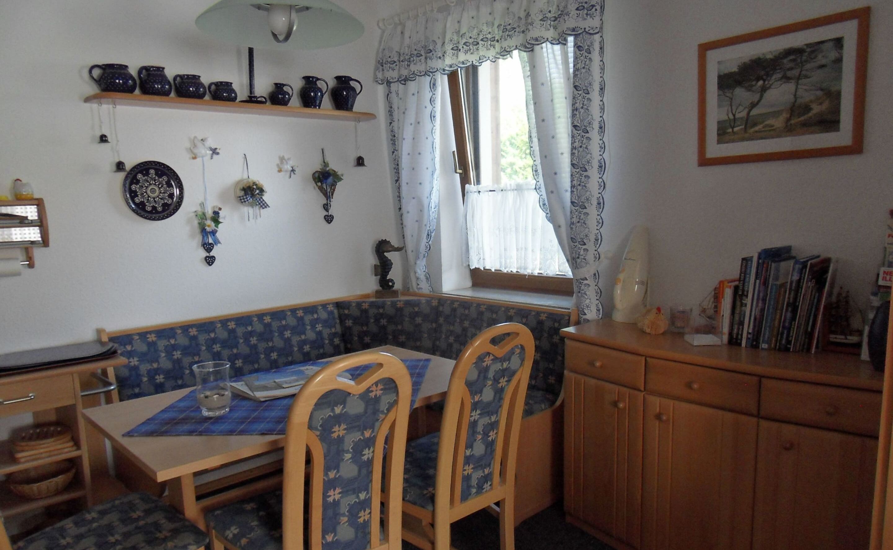 Un'accogliente sala da pranzo con una panca blu decorata, sedie in legno, un tavolo con sottopiattino e una finestra con tende di pizzo.