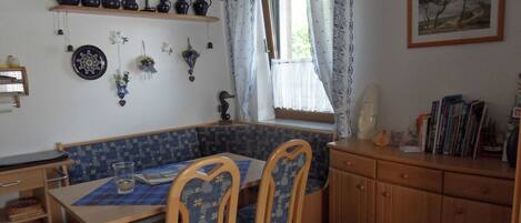 Un'accogliente sala da pranzo con una panca blu decorata, sedie in legno, un tavolo con sottopiattino e una finestra con tende di pizzo.
