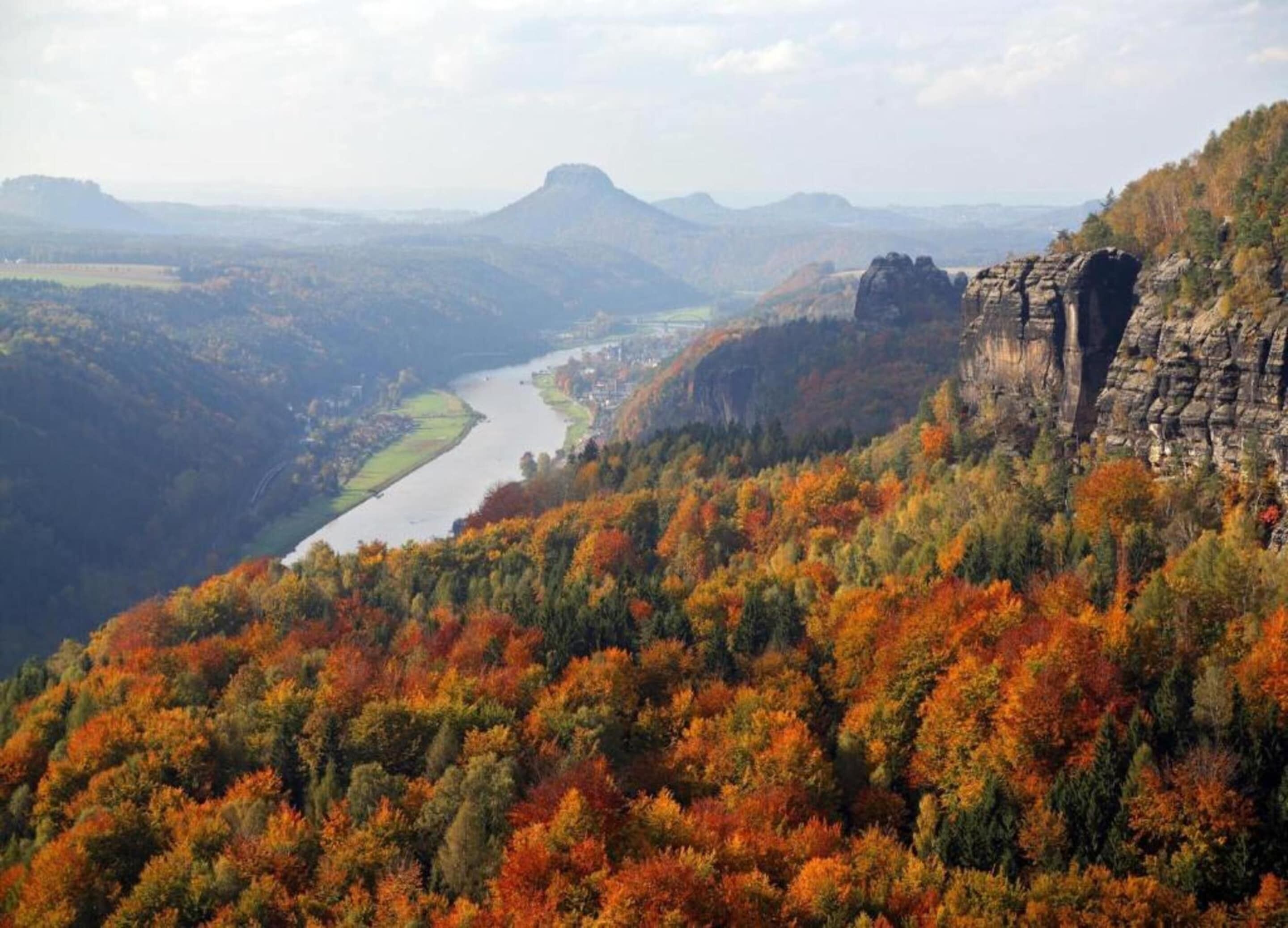Ein Fluss schlängelt sich durch ein Tal mit Herbstlaub und einer markanten Felsformation auf der rechten Seite.