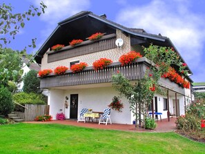 Une maison à deux étages dotée d’un balcon orné de fleurs rouges, d’un petit porche avec une table et des chaises, et d’une façade en briques.