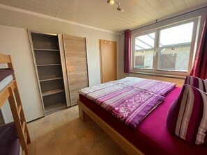 A bedroom with a bed, a wooden wardrobe, a window with curtains, and a view of the outside. - Warnkenhagen Garden Bungalow (Kalkhorst)