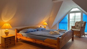 A bedroom with a sloped ceiling, a large window, a wooden bed, two bedside lamps, and a wicker chair. - Wohnung mit Balkon-nur 500 Meter zum Strand (Zingst)