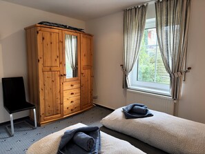 A bedroom with a wooden wardrobe, a chair, two beds, and a window with curtains. - Ferienwohnung in Zingst mit Terrasse (Zingst)