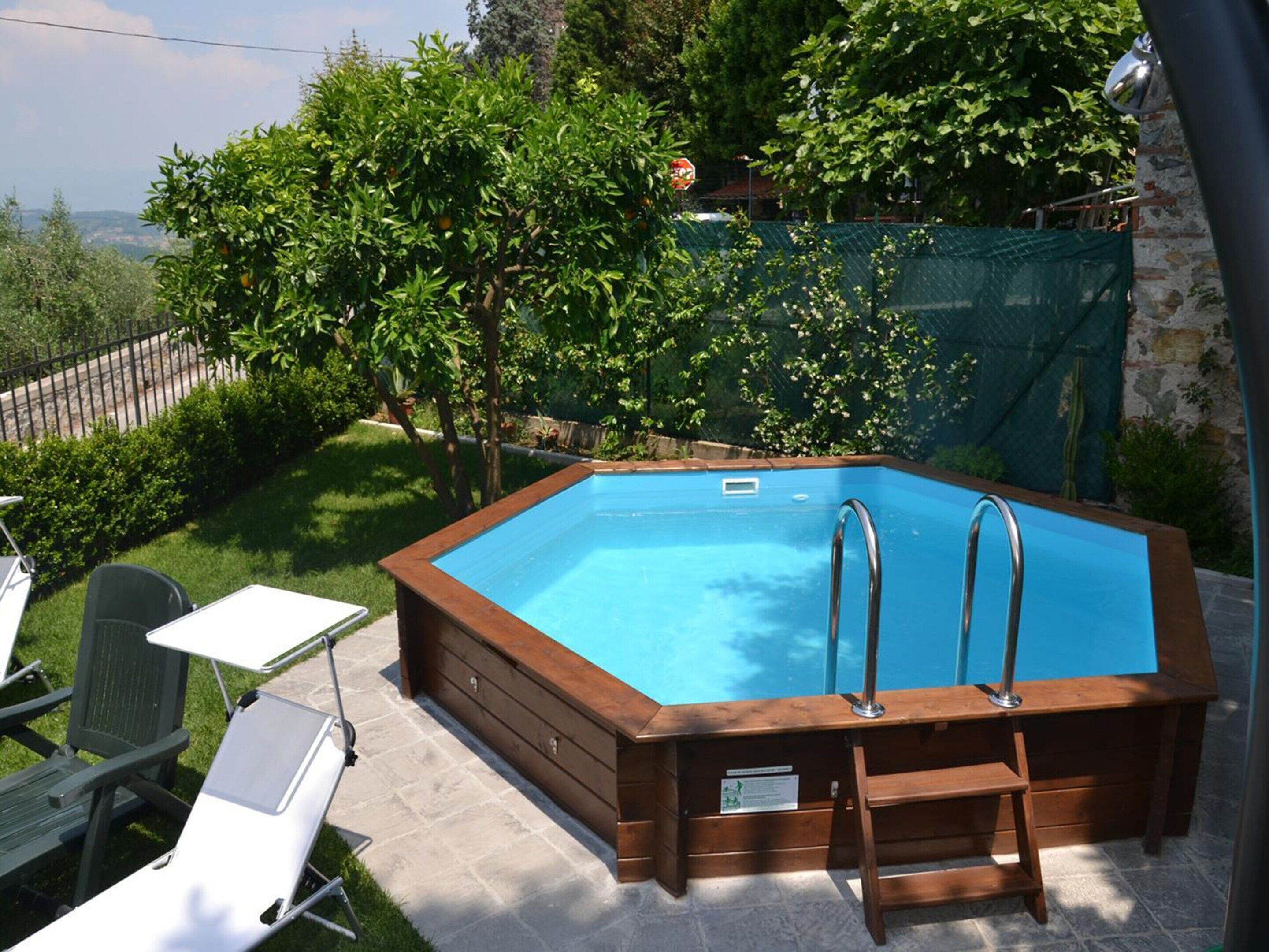 A rectangular swimming pool with a wooden deck and metal ladders, surrounded by greenery and a stone wall.