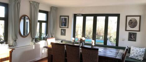 A dining area with wicker furniture, a table, and chairs. There are windows with curtains, a radiator, and framed pictures on the wall.