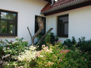 A house with two windows, a wooden sculpture, and a garden with various plants and flowers.