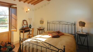 A bedroom with a wooden ceiling, a bed with a metal frame, two bedside tables with lamps, a desk with a chair, and a window with a view of greenery.