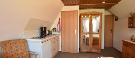Habitación pequeña con kitchenette, una cama, un armario de madera y una puerta que da a otra habitación.