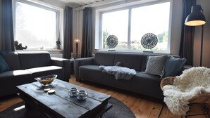 A modern living room with a grey sofa, a wooden coffee table, and a white furry chair. There are two large windows with curtains, a lamp, and decorative wall art. - Chalet in Levinska Ole Near Giant Mountains (Levínská Olešnice)
