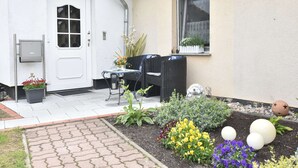 A residential exterior with a white door, a window with a curtain, a garden with various plants, and outdoor furniture. - Moderne Ferienwohnung mit Grill in Rakow (Am Salzhaff)