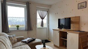 A cozy living room with a sofa, a small table, a TV on a wooden stand, and a window with curtains. - Charmante Ferienwohnung mit Garten in Wiek (Wiek)