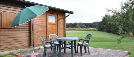 A wooden cabin with a patio area featuring a table and chairs, a green umbrella, and a brick pathway.