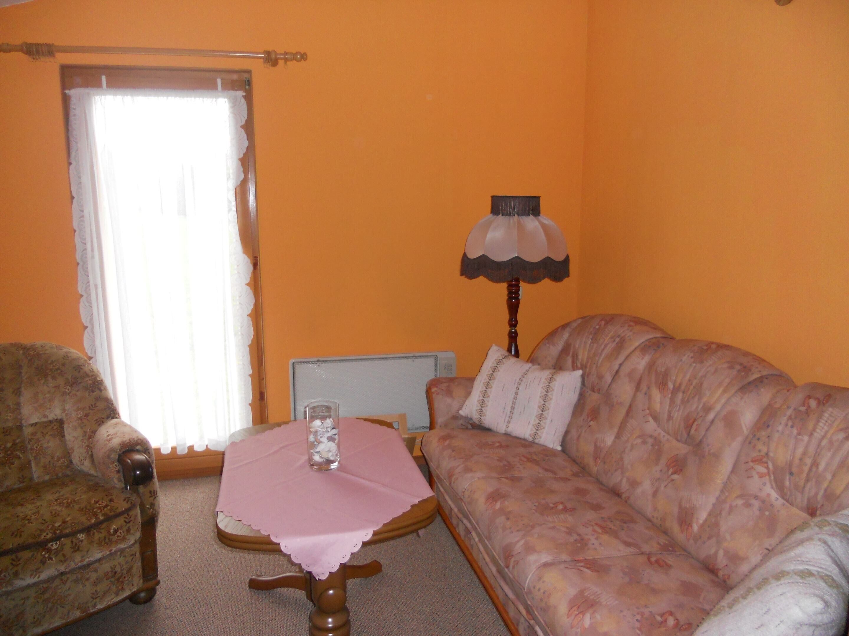 Ein Wohnzimmer mit einem blumengedeckten Sofa, einem Holz-Sessel, einem kleinen runden Tisch mit rosa Tischdecke und einer Lampe.