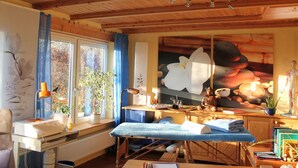 Ein Holzraum mit einer Massageliege, einem Fenster mit blauen Vorhängen, einer Topfpflanze und einem Gemälde von Blumen und Steinen.