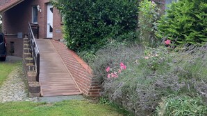 Ein Backsteinhaus mit einer Holzrampe, die zu einer weißen Tür führt, umgeben von üppigem Grün und blühenden Pflanzen.