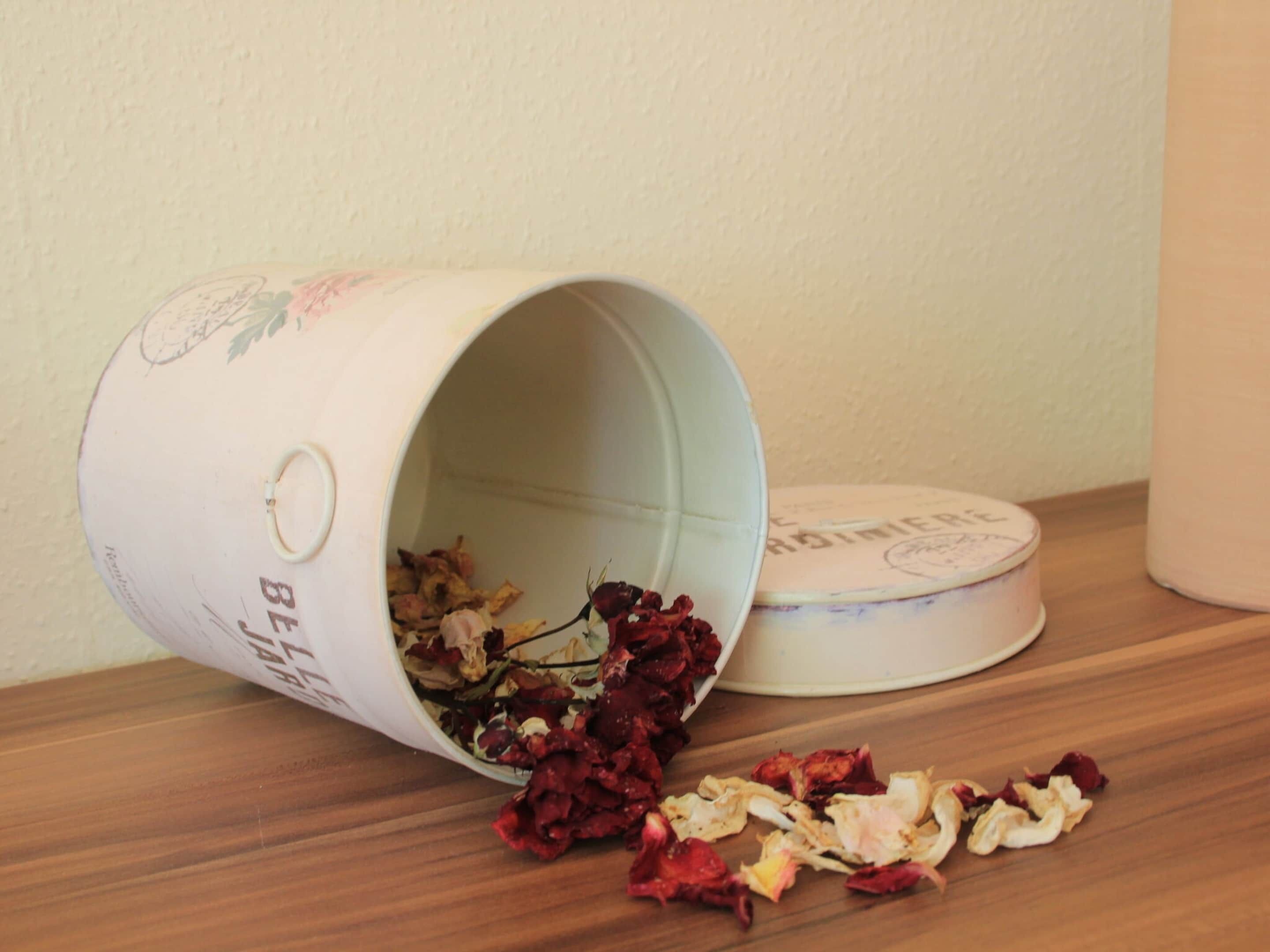 A white ceramic container with floral designs and text, tipped over with dried flowers spilling out onto a wooden surface.