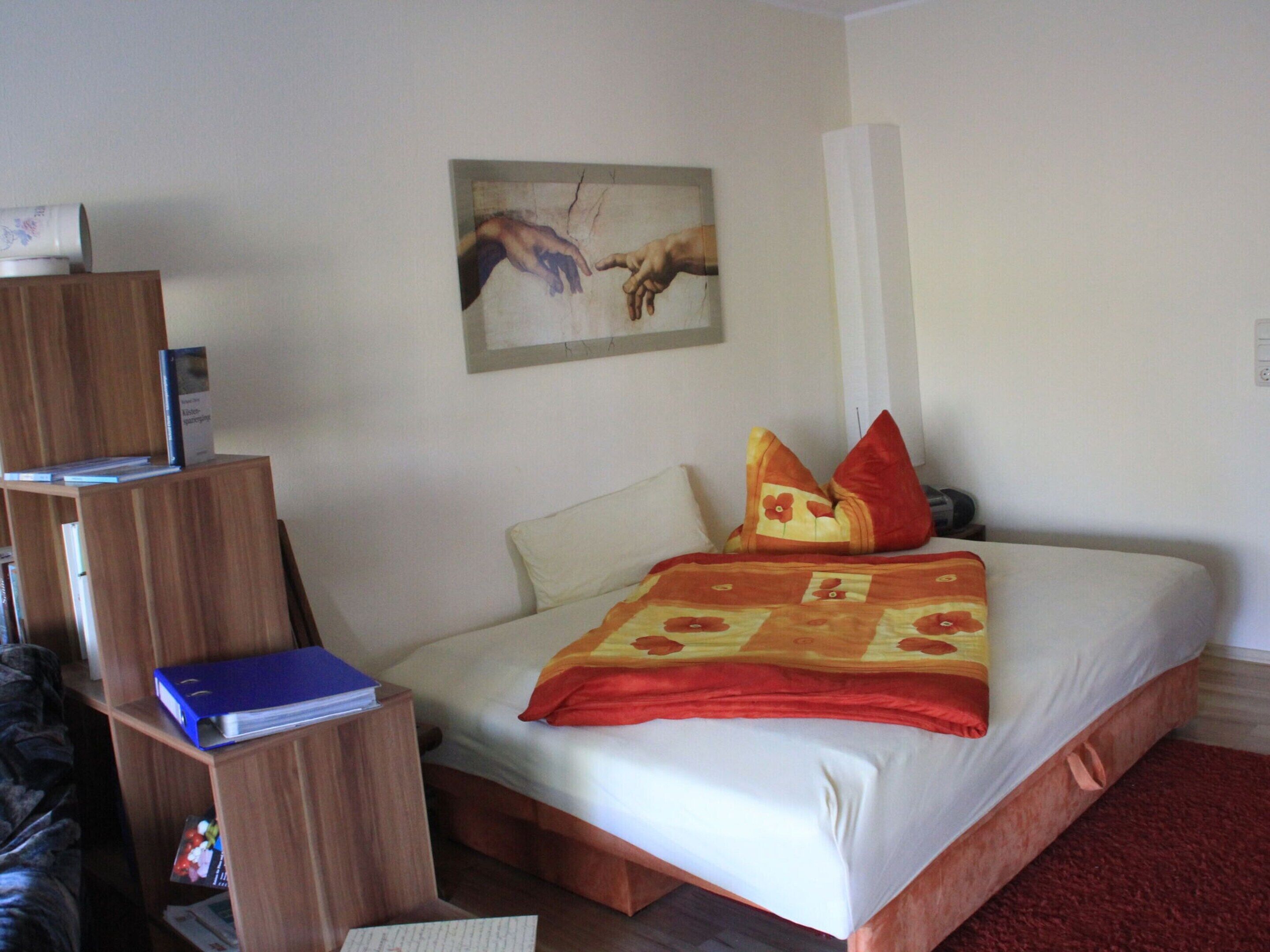 A hotel room with a bed, a wooden desk, a bookshelf, and a painting on the wall.
