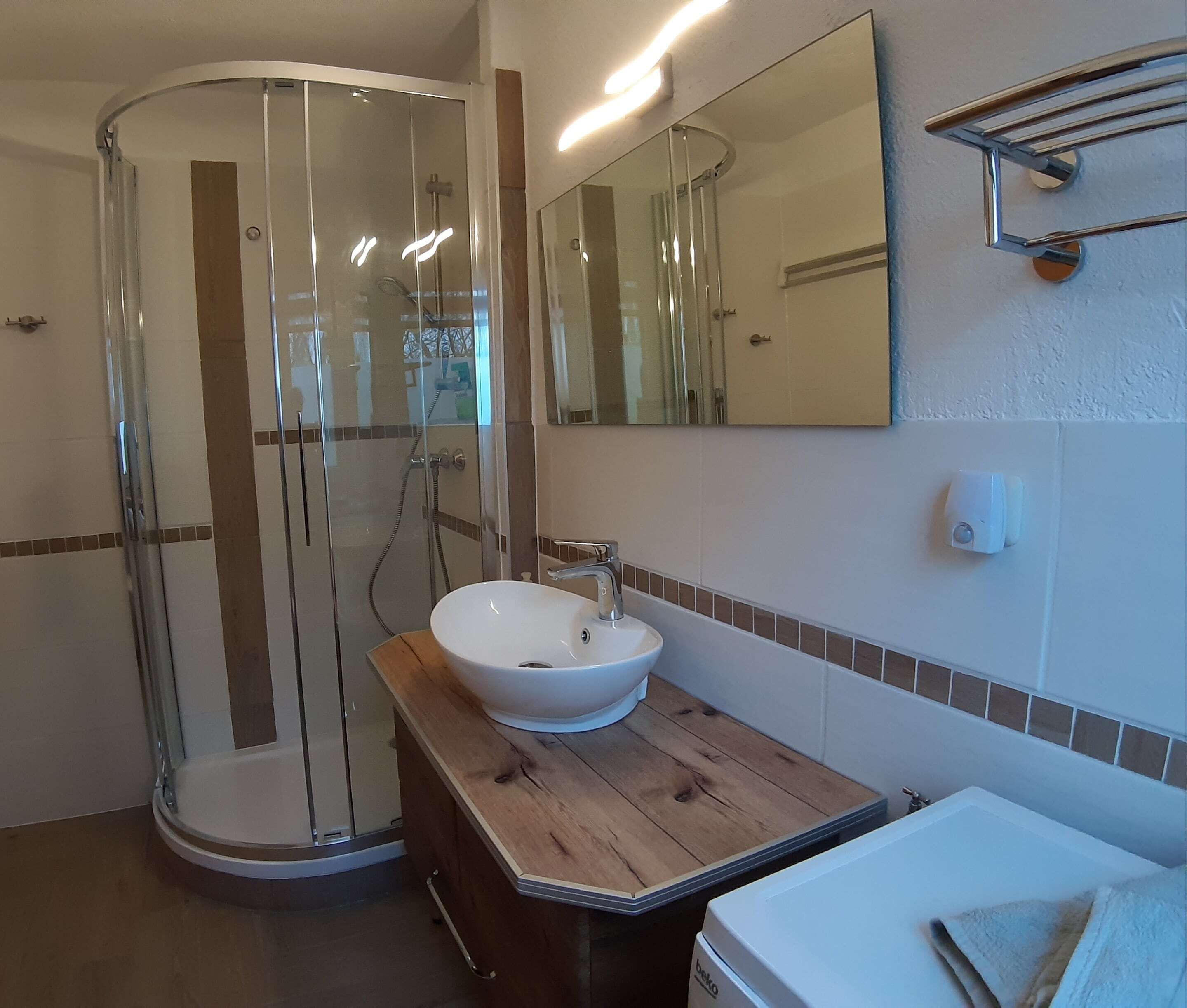 A modern bathroom with a glass-enclosed shower, a white sink with a wooden countertop, and a large mirror above the sink.