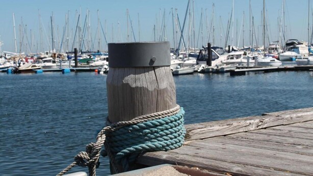 A wooden dock with a rope tied to a post, a marina with multiple sailboats, and a clear blue sky.
