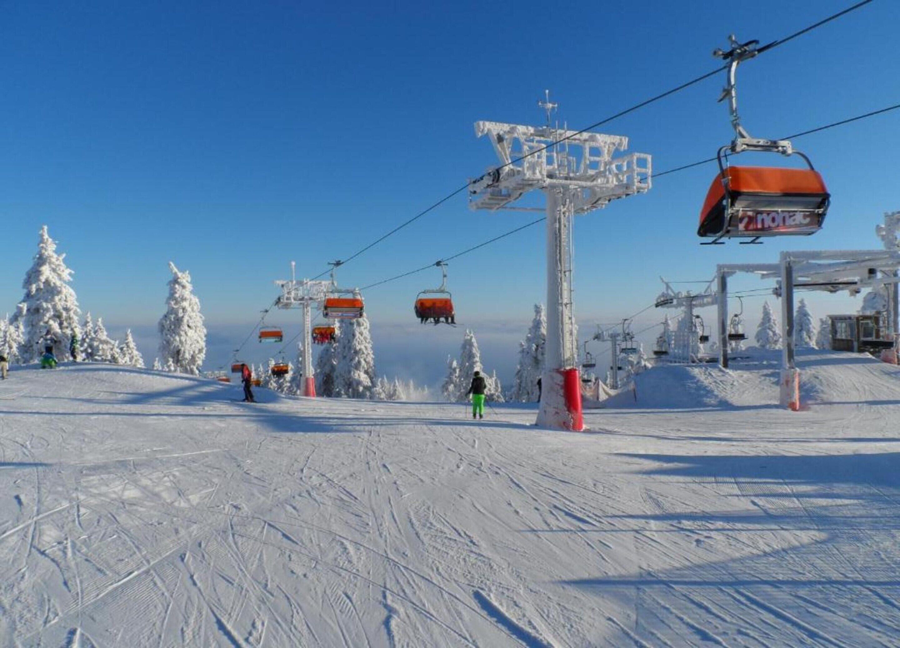 Gondola de esqui com cadeiras e placa, rodeada por árvores cobertas de neve e um céu limpo.