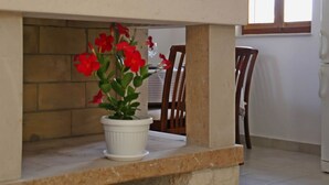 Una maceta con flores rojas sobre un alféizar de piedra en una habitación con suelo de baldosas y una ventana con persianas.